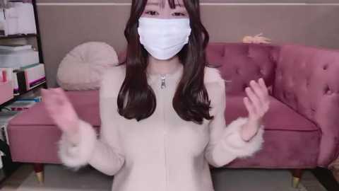 Media: Video of an Asian woman with long brown hair, wearing a white face mask, white fur-trimmed coat, and a silver necklace, gesturing with her hands in front of a pink velvet couch.