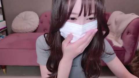 Media: Video of a young East Asian woman with long, dark hair and pale skin, wearing a gray t-shirt and a white face mask, sitting on a pink velvet sofa, holding her nose, indoors.