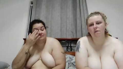 Media: Video of two plus-size, topless women with fair skin and medium-length hair, one with a tattoo on her arm, sitting on a bed with a gray curtain behind them.