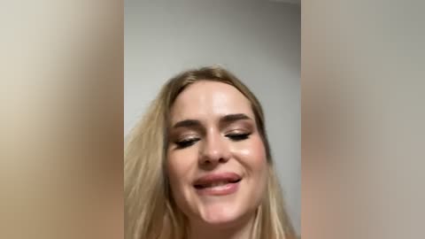 Video of a smiling, fair-skinned blonde woman with light makeup, slightly blurred in the background. She has long, straight hair and is wearing a subtle, neutral outfit.