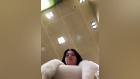 Media: A video captures a woman with dark hair, wearing a white fur coat, standing in a beige-tiled bathroom with recessed lighting and a green wall. The perspective is from a low angle, emphasizing her height and the room's layout.