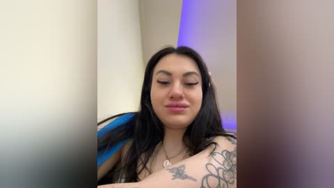 Media: Video of a young woman with long black hair, light skin, and a serene expression. She wears a blue and white striped shirt, showing tattoos on her arm. Background is a blurred beige and purple gradient.