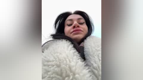 Media: Video of a woman with long black hair, eyes closed, wearing a fluffy white coat, against a blurred background.