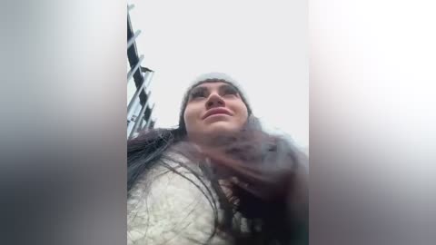 A video of a young woman with long dark hair, wearing a gray beanie and white scarf, smiling from a low angle, with blurred background objects.