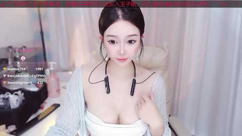 Media: Video of a young East Asian woman with light skin, dark hair in a bun, wearing a white off-shoulder top, black headphones, and a light cardigan. Background shows a white curtain and a desk with a black camera.