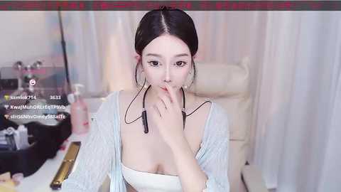 Media: A video of an East Asian woman with light skin and black hair tied back, wearing a white top and a light blue cardigan, sitting at a desk with a water bottle and a headset, looking serious.