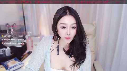 Media: Video of an East Asian woman with long black hair and fair skin, wearing a light blue cardigan, sitting indoors with makeup and hair products on a table.