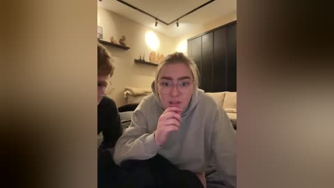 Media: A candid video shows a woman with glasses, wearing a beige sweater, leaning over a table in a dimly lit living room, with a mirror and shelves in the background.