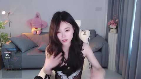 Media: Video of an East Asian woman with long black hair, wearing a black lace dress, taking a selfie in a modern living room with a gray sofa, plush toys, and a floor lamp.