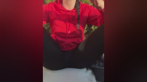 Media: Video of a person in a red hoodie, black leggings, and white sneakers, sitting with legs spread apart, revealing black underwear, against a blurred green background.