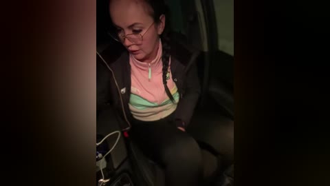 Media: A video shows a woman with glasses, wearing a pink and green striped shirt and black jacket, leaning over a cup in a dimly lit car.