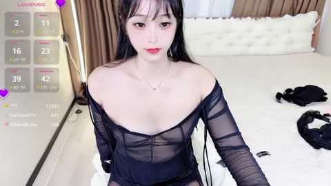 Media: A young Asian woman with pale skin, long black hair, and red lipstick sits on a bed in a modern bedroom, wearing a sheer black dress, revealing her cleavage.