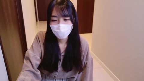 Media: A video of an Asian woman with long black hair, wearing a beige robe and a white mask, leaning against a wall in a dimly lit, narrow hallway with white floors and dark wooden doors.