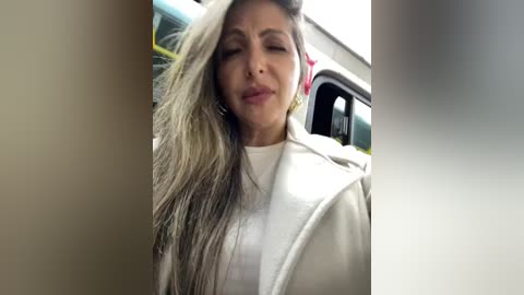 Media: A video of a woman with long, wavy, gray-streaked hair, wearing a white hoodie, smiling slightly, standing outdoors next to a bus.