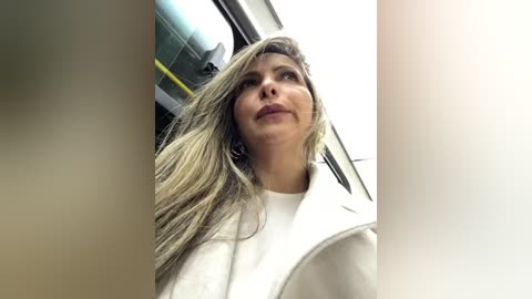 Media: A video of a blonde woman with long hair and fair skin, wearing a white top, captured from a low angle in a modern, bright indoor setting.