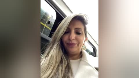 Media: A video of a woman with long, straight blonde hair, wearing a white top, smiling inside a car, with blurred vehicle interior and daylight.