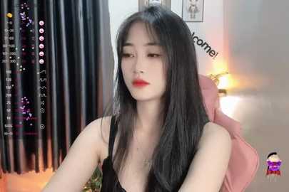 Media: A video of an Asian woman with long black hair and fair skin, wearing a black tank top, sitting on a pink chair in a dimly lit room.