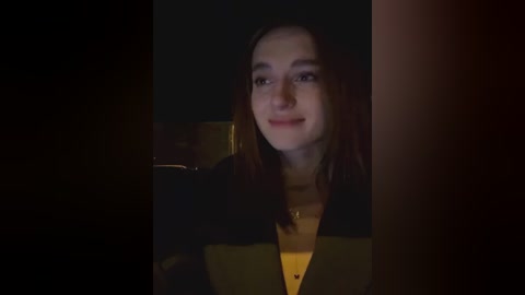 Media: Video of a young woman with fair skin and straight, shoulder-length brown hair, smiling slightly, wearing a black jacket over a yellow top. The dimly lit background is indistinct.