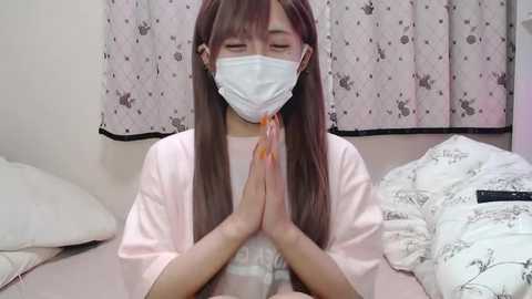 Media: A video of an Asian woman with long brown hair, wearing a white mask, pink shirt, and closed eyes, praying with hands clasped together, in a room with white curtains and floral-patterned bedding.