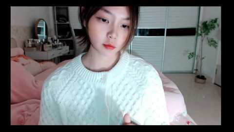 Media: A young Asian woman with pale skin, dark hair, and a large nose ring, wearing a white knitted sweater, sits on a pink bed in a brightly lit bedroom.