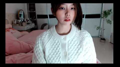 Media: Video of an Asian woman with straight black hair, wearing a white knitted cardigan, sitting on a bed with pink sheets in a softly lit bedroom.