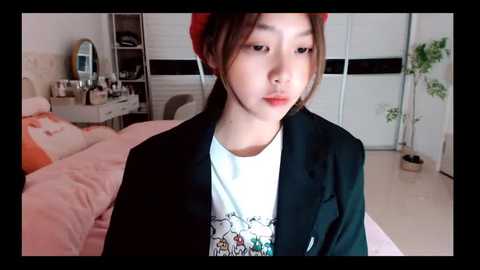 Media: Video of a young Asian woman with fair skin, wearing a black blazer over a graphic tee, sitting in a minimalist bedroom with pink bed, white wall, and plants.