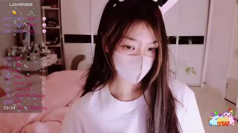 Media: A video of an East Asian woman with long, straight black hair wearing a white mask and bunny ears, standing in a bedroom with pink bedding and white walls.