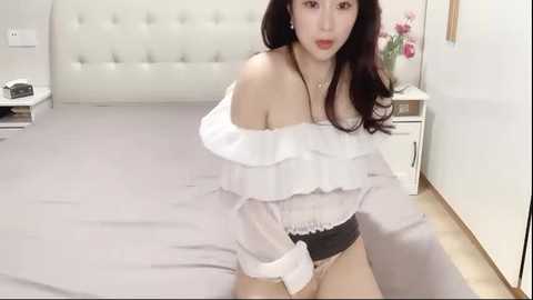 Media: Video of an Asian woman with long, dark hair, wearing a white off-shoulder ruffled blouse, black panties, and sitting on a bed in a modern, minimalist bedroom with white walls and a tufted headboard.