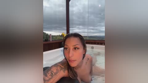 Media: Video of a nude woman with wet hair, medium breasts, and tattoos, lounging in a bathtub with a scenic outdoor view.