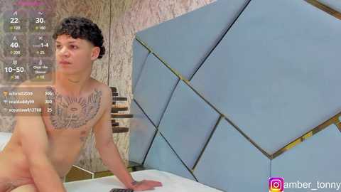 Media: A young, shirtless, curly-haired, light-skinned man with tattoos sits on a bed with a blue, geometric-patterned headboard in a modern, beige-tiled room. The image includes a timestamp and social media icons.