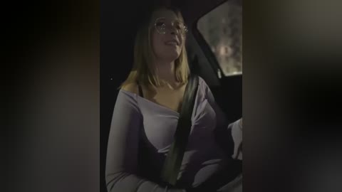 Media: Video of a blonde woman in glasses, wearing a purple off-shoulder top, driving at night with dim lighting. She appears calm and focused, with a safety belt across her chest.