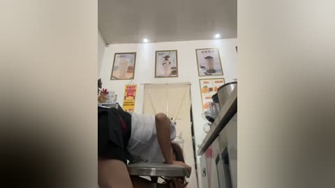 Media: Video of a man in black shorts and a white shirt, leaning over a kitchen counter. The background features framed posters, a sink, and a microwave.