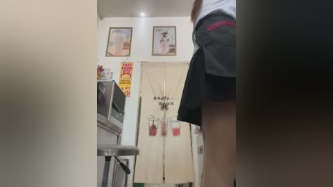 Media: Video of a woman in a short black skirt and white top, standing in a small, cluttered laundry room with white walls, framed photos, and laundry machines.