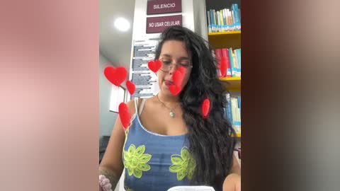 Media: Video of a young Latina woman with long black hair, wearing a blue tank top with green floral print, seated in a library, heart emojis overlaying her face.