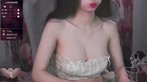 Media: A video of a young Asian woman with long black hair, wearing a white lace bra, sitting on a bed. The background is blurred with a dark color scheme and floral decor.