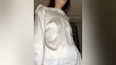 Media: Video of a woman with light skin, wearing a white, long-sleeved blouse, partially visible face, indoors with blurred background featuring a doorway and beige walls.