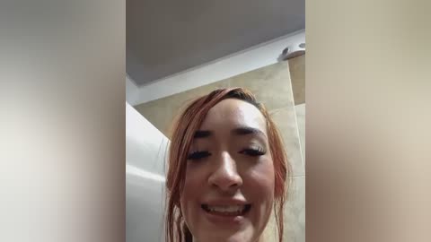 Media: A close-up video of a smiling woman with long, straight, light brown hair, captured from a low angle. The background features a beige tiled wall and a white ceiling, suggesting a bathroom setting.