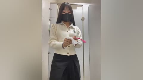 Media: A video of an Asian woman with long black hair and a black face mask, wearing a white cardigan and black skirt, standing in a hallway.