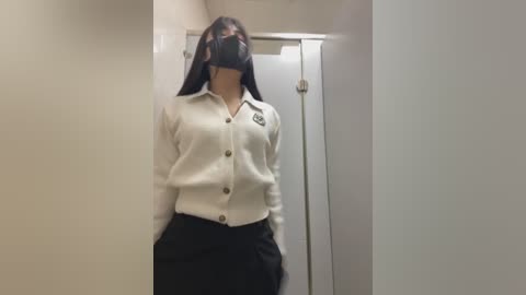 Media: A video of a woman in a white cardigan, black mask, and black skirt, standing in a narrow, dimly-lit hallway with beige walls and a closed door.