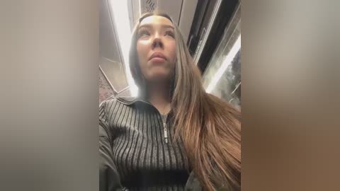 Media: Video of a young woman with long, straight brown hair, wearing a black, ribbed, long-sleeved top, standing inside a dimly lit, narrow airplane aisle.