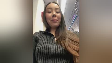 Media: Video of an Asian woman with long, straight, brown hair, wearing a black ribbed sweater, standing in a public restroom with metal towel racks and a hand dryer in the background.