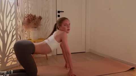 Media: Video of a fit, light-skinned woman with long blonde hair in a white sports bra and black leggings, performing a plank on a light wooden floor in a minimalist room with beige walls and a closed door.