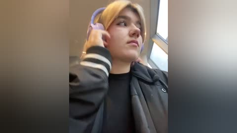 Media: A video of a young Caucasian woman with blonde hair, wearing a black jacket, adjusting headphones, looking contemplative by a window in a dimly lit room.