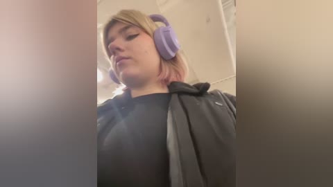 Media: A low-angle video of a young woman with light skin and short blonde hair, wearing large purple headphones and a black hoodie, standing in a dimly lit room with white walls.