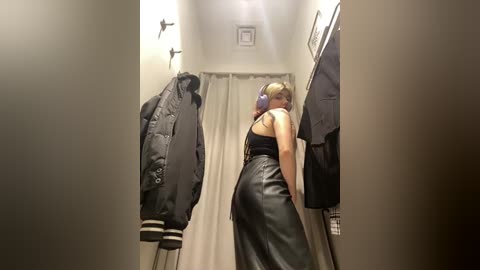 Media: A video of a woman in a dressing room, wearing a black leather skirt, blue headphones, and a sleeveless top, looking at clothes on a rack.