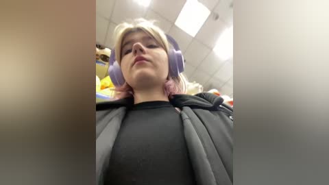 Media: A low-angle video of a young woman with light skin, shoulder-length blonde hair, wearing purple headphones, a black hoodie, and a gray jacket. She stands in a brightly lit room with white ceiling tiles and a yellow toy in the background.