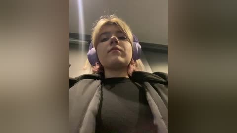 Media: Video of a young woman with fair skin, blonde hair, wearing large purple headphones and a black hoodie, lying on a dimly lit floor with blurred walls.