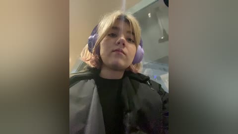 Media: Video of a young woman with blonde hair, wearing purple headphones, a black top, and a grey jacket, sitting indoors, blurry background.