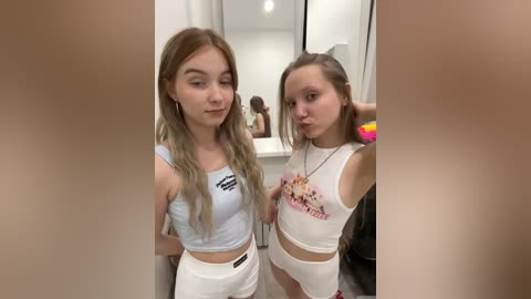 Media: Video of two young women with light skin and long blonde hair, wearing matching white crop tops and pants, posing in a modern bathroom with mirrors and bright lighting.