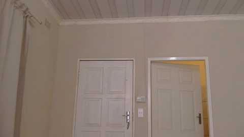 Media: Video of a plain, beige-walled room with a white, paneled door and silver doorknob, an open doorway revealing a warm-toned interior, and a white ceiling with horizontal grooves.